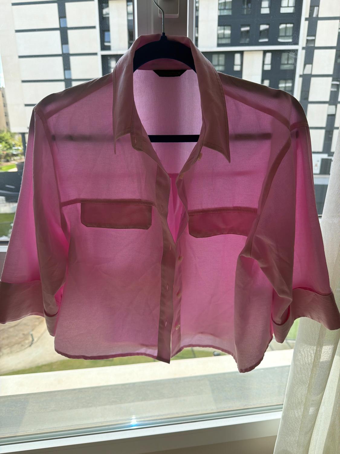 Translucent pink shirt hanging on a hanger in front of a window, with buildings in the background and natural light illuminating it.