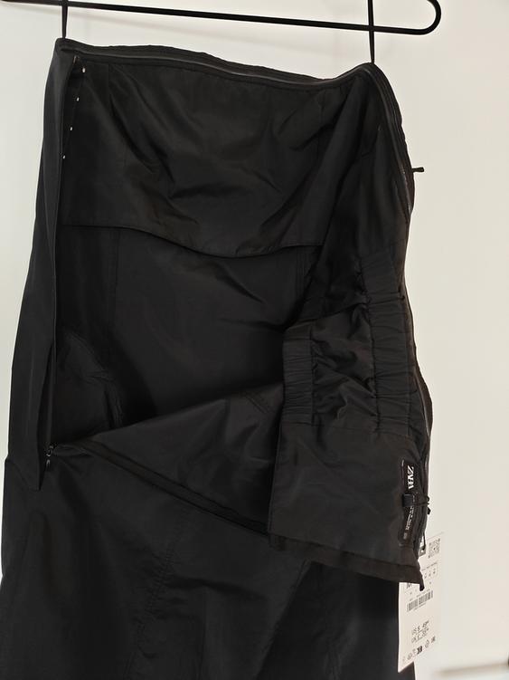 Black trousers hanging on a hanger, with a visible label that says "ZARA". Fabric with pleats and a side zip. White background.