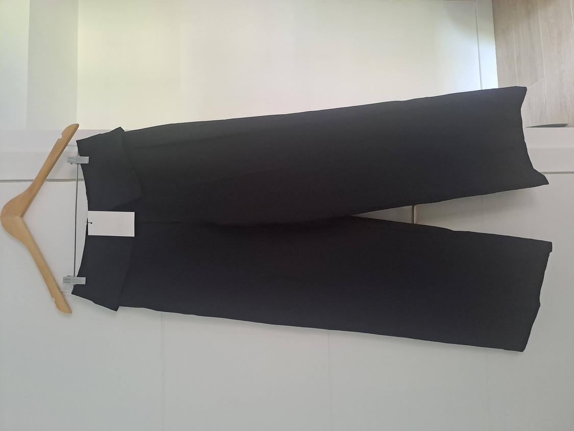 Black wide-leg trousers hanging on a wooden hanger with clips against a white background. They have a folded waist and a tag.