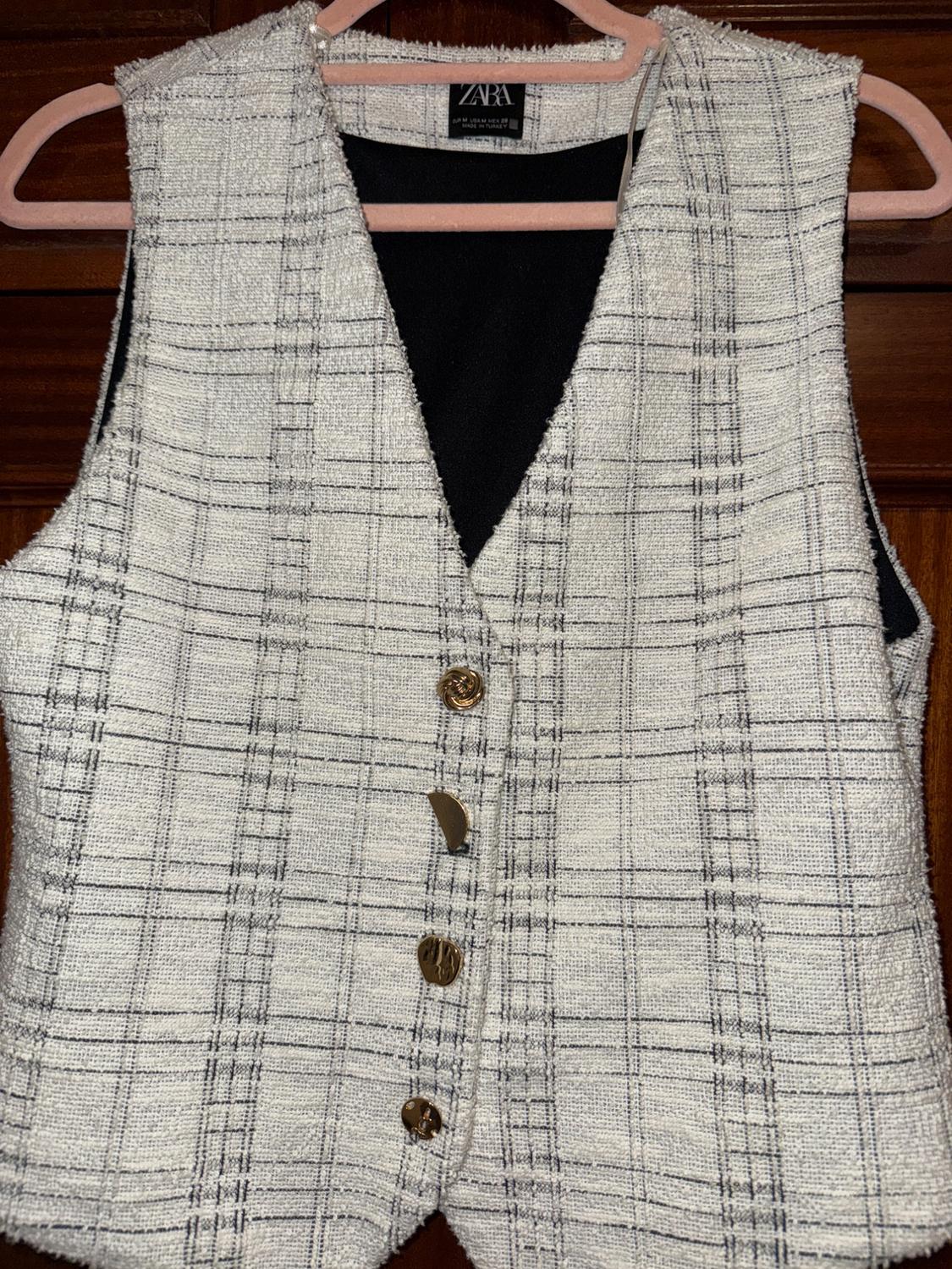 Sleeveless waistcoat with a black and white checked print, gold buttons, and Zara label, hanging on a pink hanger.
