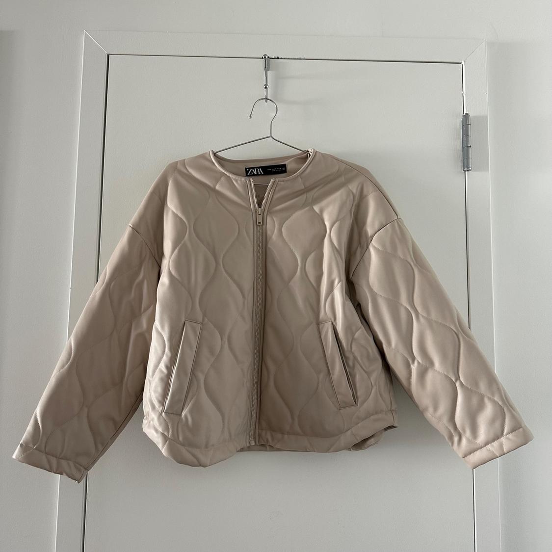 Beige puffer jacket hanging on a hanger over a white door. Design with front zipper and side pockets.