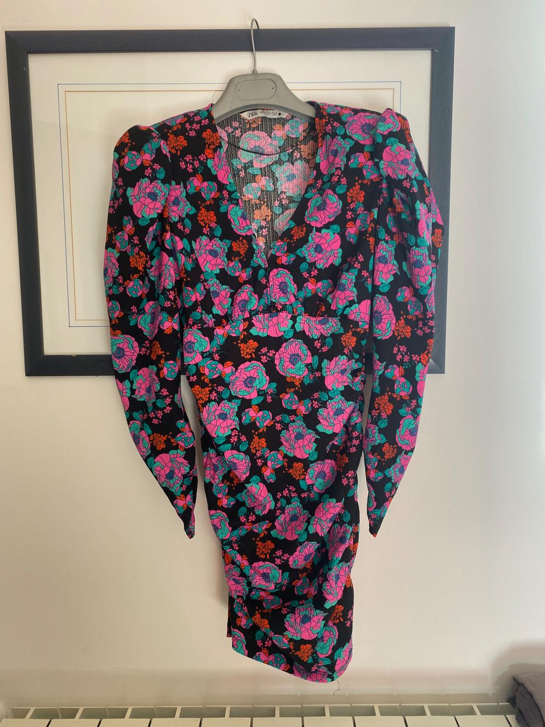 Long-sleeved floral dress with a black background, printed with pink, green, and orange flowers, hanging on a hanger in front of a painting.