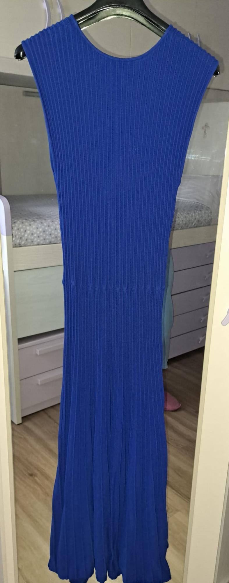 Long blue ribbed sleeveless dress hanging on a hanger in front of a mirror reflecting a bedroom with a bed and wooden floor.