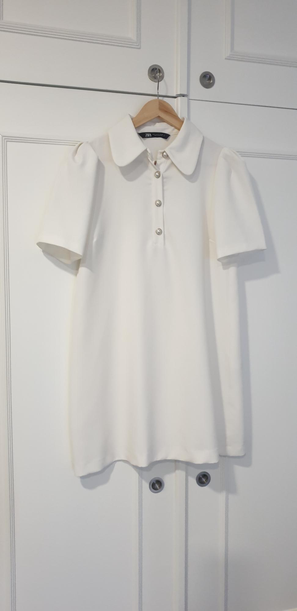 White short-sleeved dress with a round neck and buttons down the front, hanging on a wooden hanger in front of a white wardrobe.