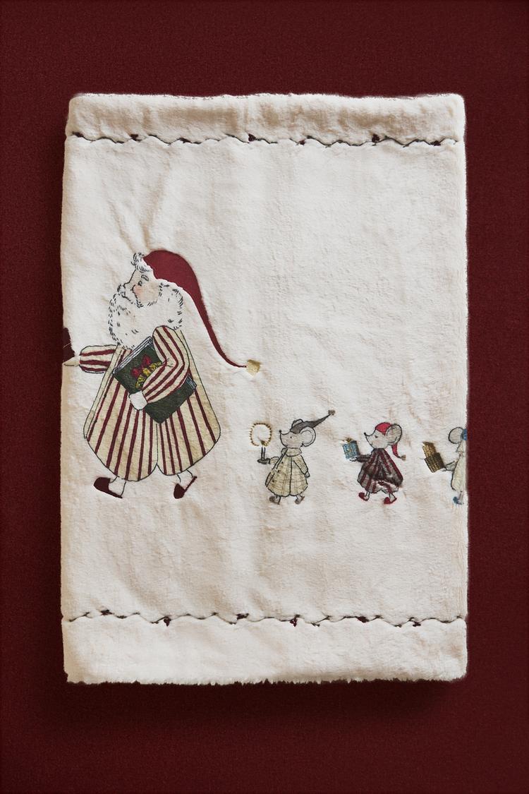 Detalj bijele deke s izvezenim Djedom Božićnjakom i miševima u nizu, s tamnim ukrasnim rubovima.