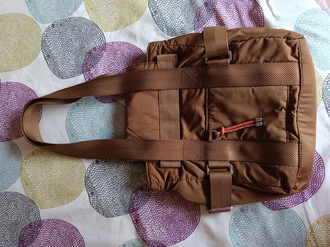 Brown handbag with straps on a bedspread with colourful circles. Red and black drawstring detail on the front pocket.