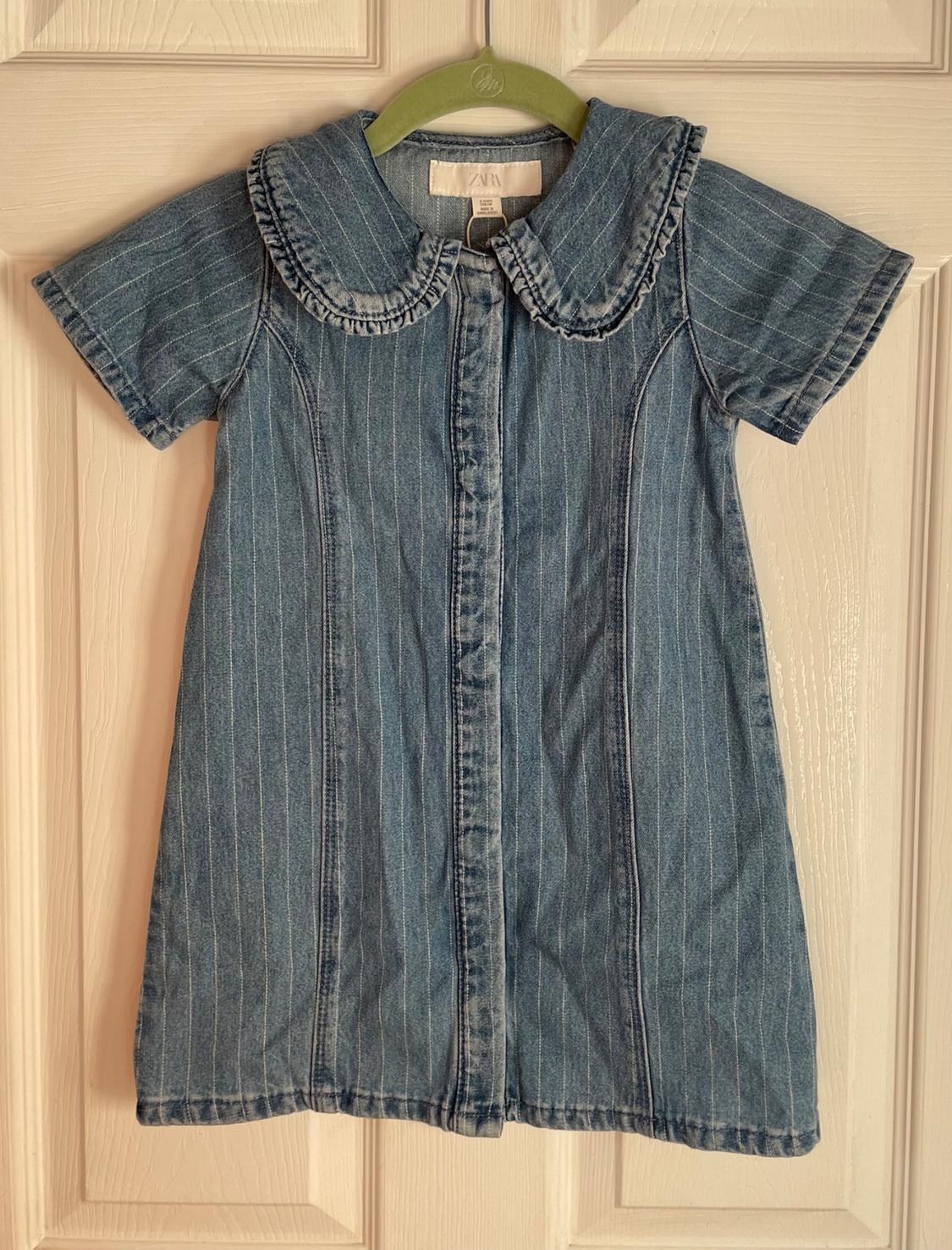 Blue denim dress with fine white stripes, round frilled neck and short sleeves, hanging on a green hanger.