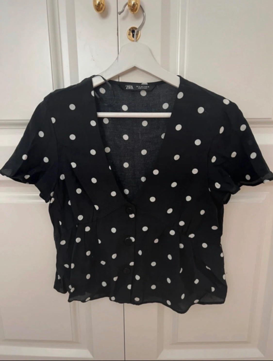 Black blouse with white polka dots, short sleeves and buttons. Hanging on a coat hanger on a white door with gold doorknobs. Text: ZARA.
