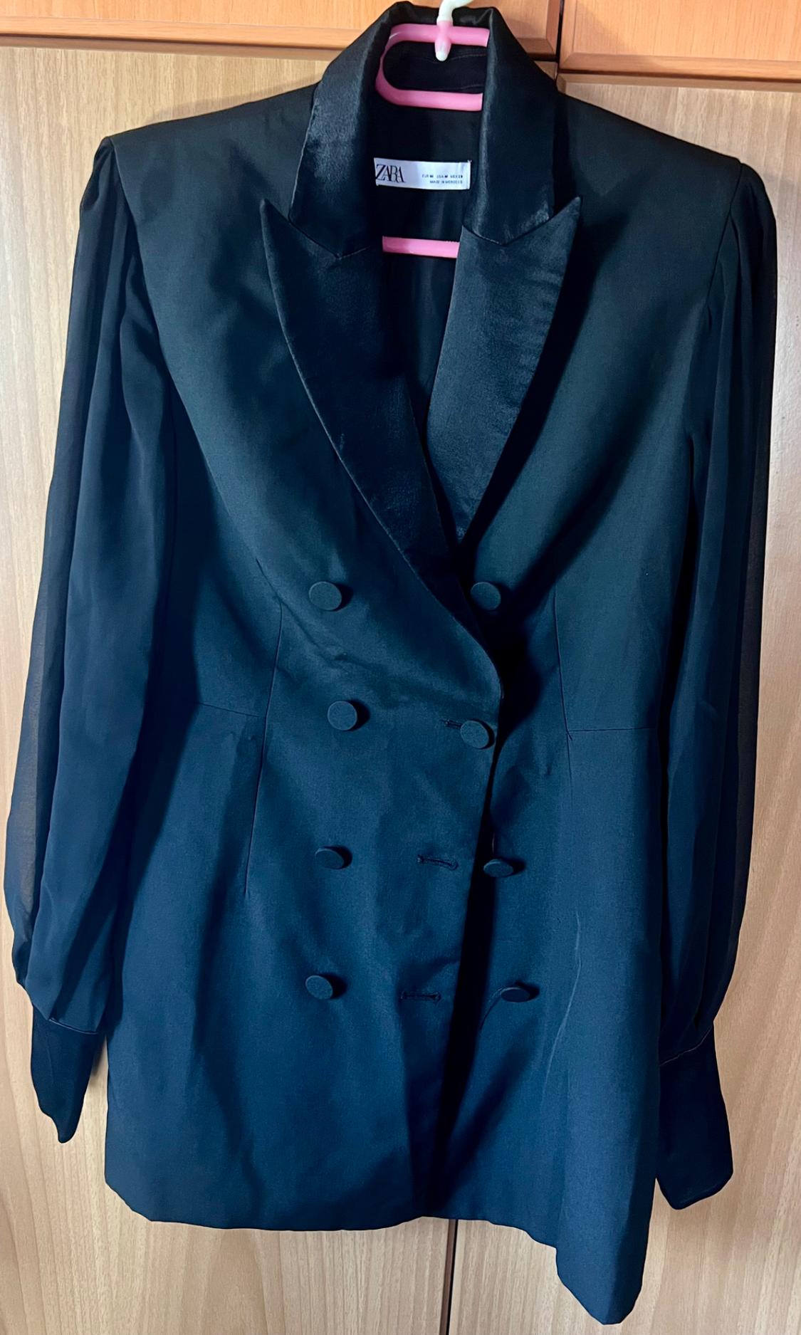 Black double-breasted blazer with ZARA label, satin lapels, semi-transparent sleeves and buttons, on a pink hanger against a wooden background.