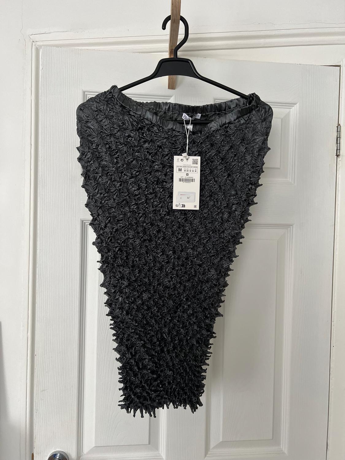 Black trousers with a crinkled texture hanging on a hanger in front of a white door. Visible tag with product details.