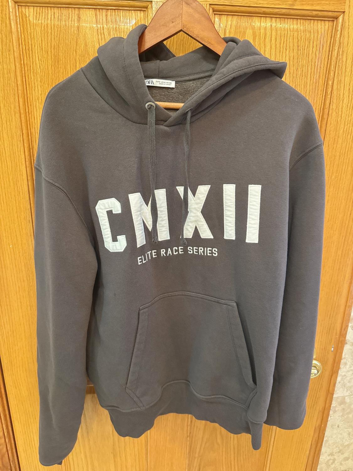 Grey hooded sweatshirt hanging on a wooden hanger. Text on the front: "CMXII ELITE RACE SERIES". Wooden door background.