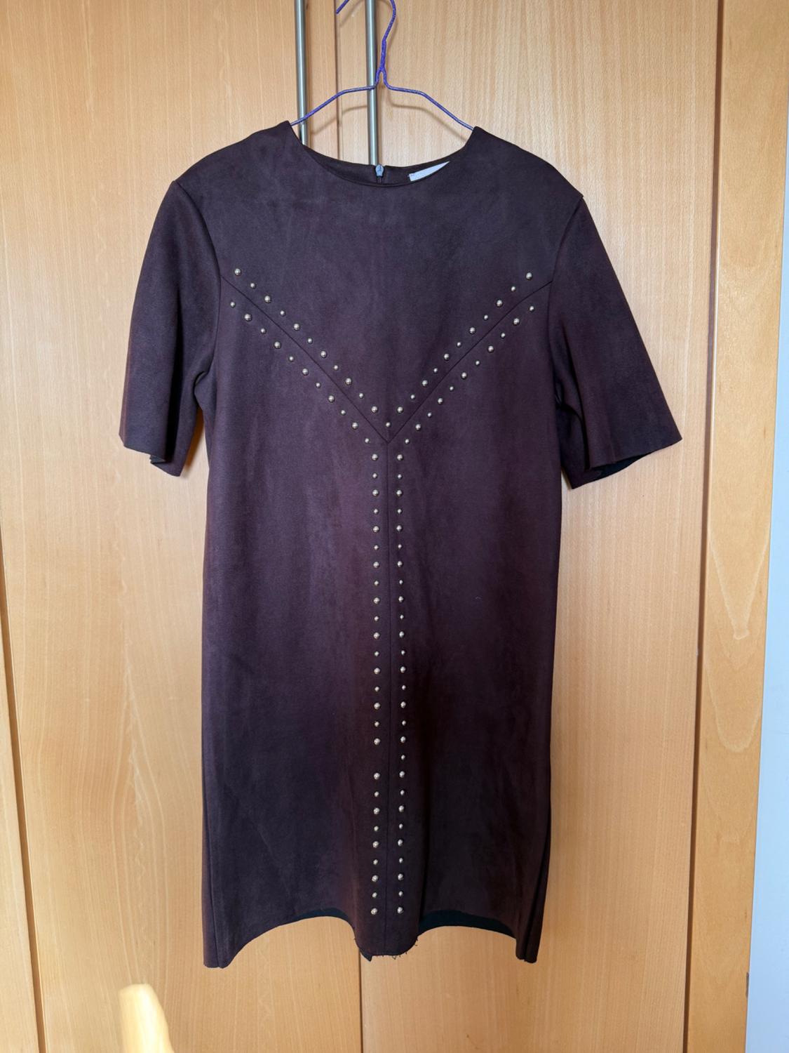 Dark brown dress with V-shaped studs on the chest and down the centre, hanging on a blue hanger against a wooden background.