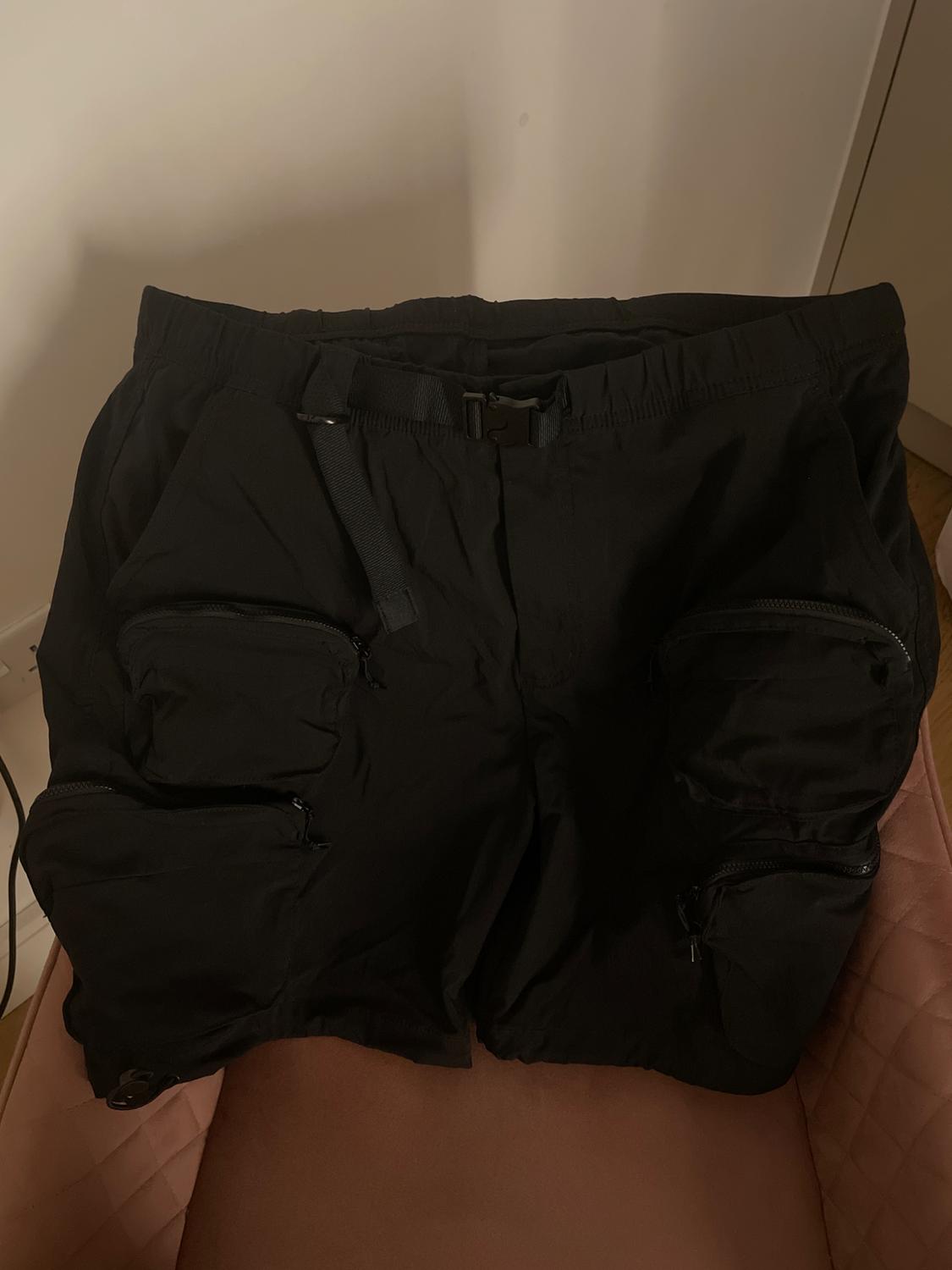Black shorts with multiple pockets and zips, placed on a pink chair in an indoor setting.