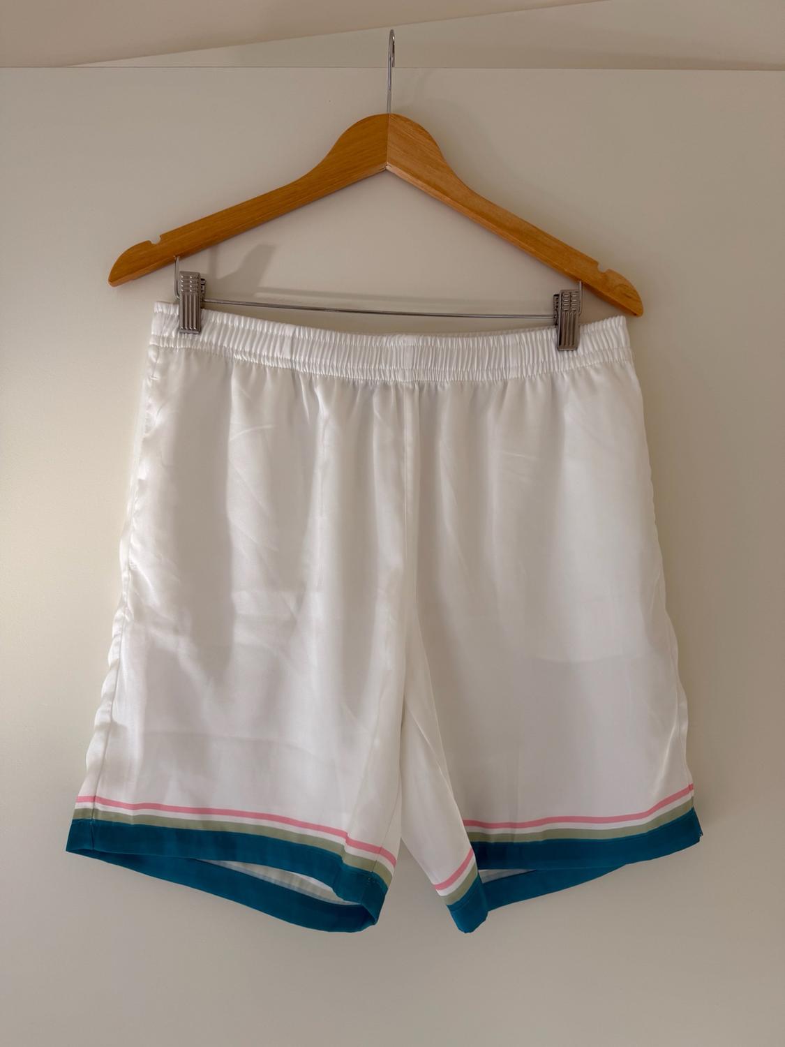 White shorts with an elasticated waist and pink, green, and blue stripes on the hem, hanging on a wooden hanger against a light background.