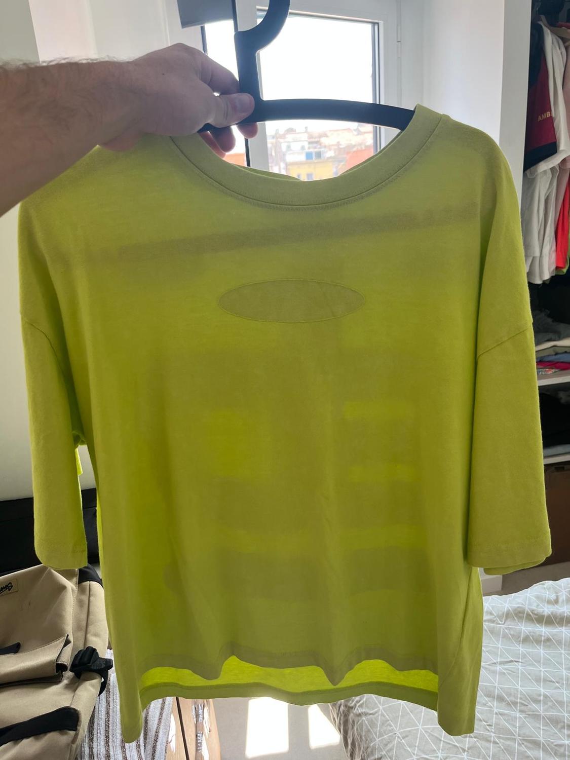 A semi-sheer yellow T-shirt is hanging on a black hanger, held by a hand in front of a window lit with natural light.
