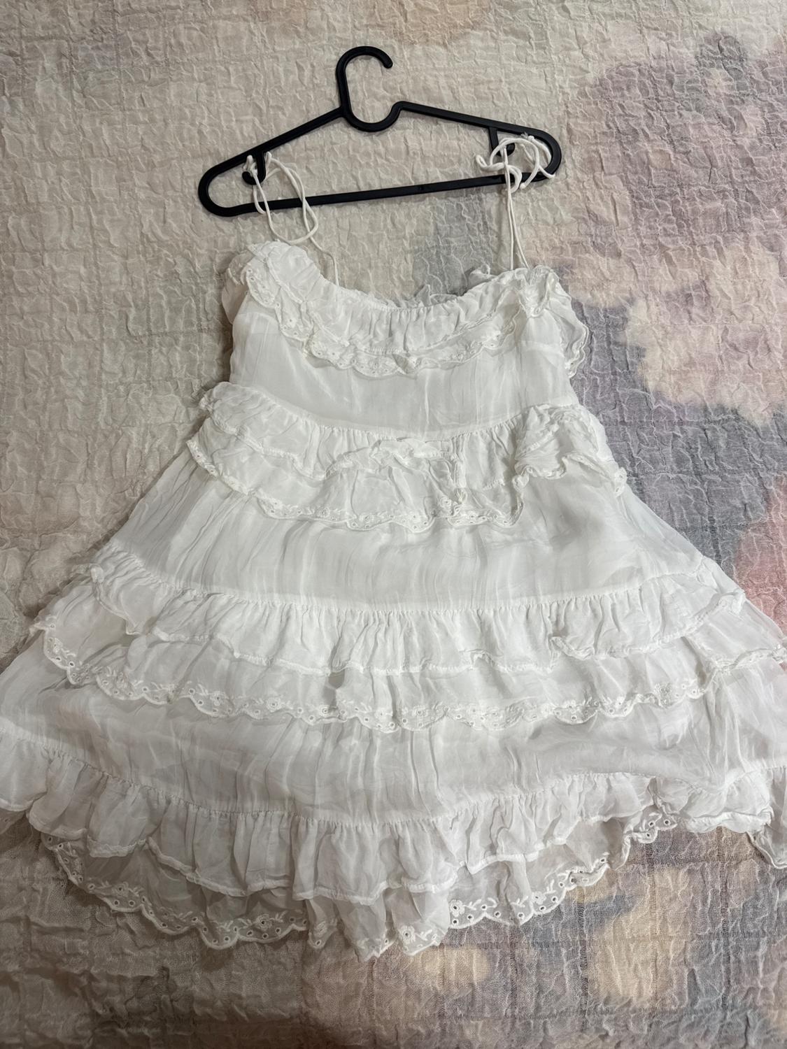 White dress with knotted straps and tiered embroidered ruffles, hanging on a black hanger over a soft-toned bedspread.