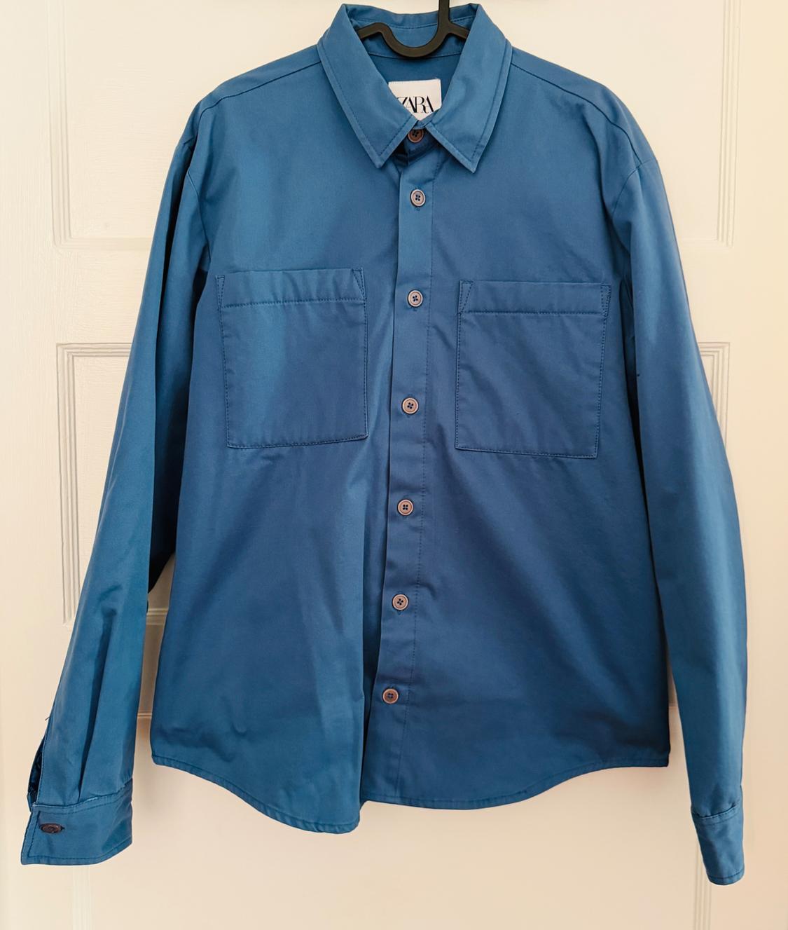 Blue long-sleeved button-down shirt with two chest pockets, hanging on a black hanger over a white door.