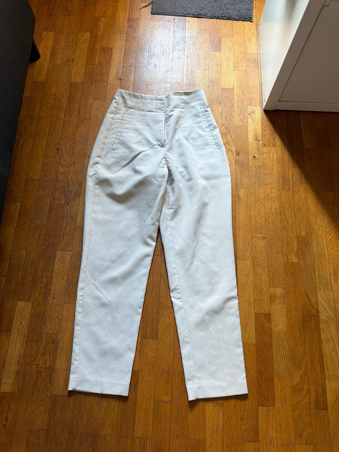 White high-waisted trousers with pleats and pockets, laid out on a wooden floor in natural light.