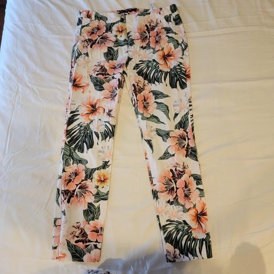 White fabric pants with a floral print in pink, orange, and green tones, on a bed with white sheets.