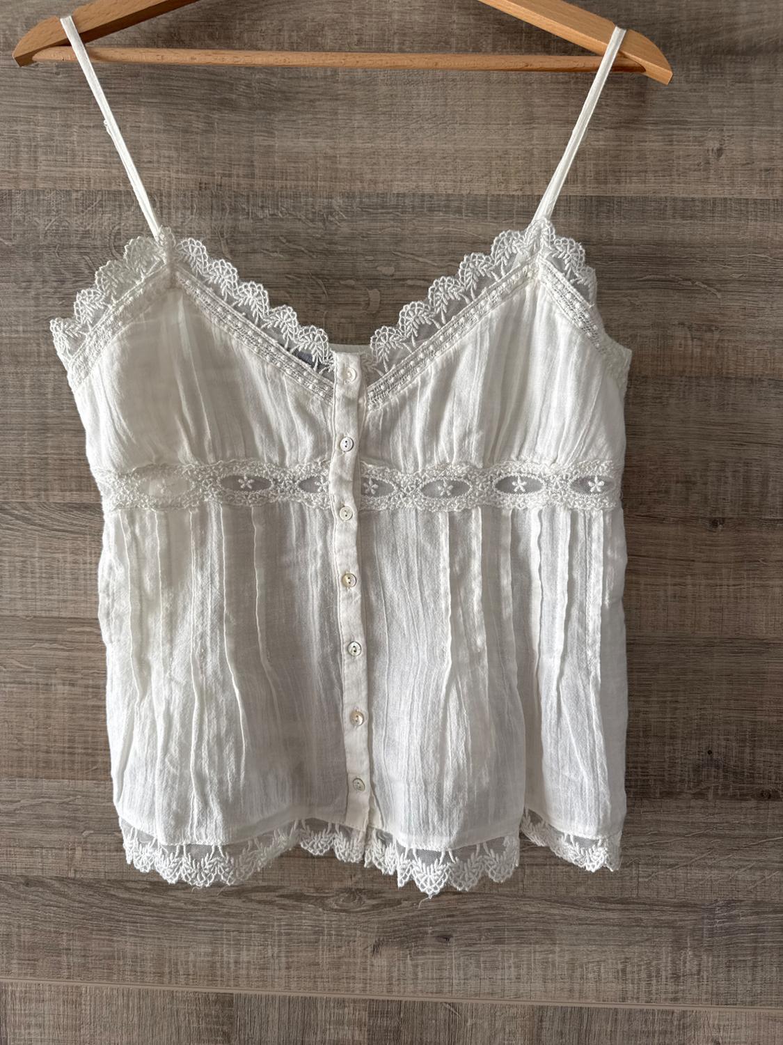 White strappy blouse with front buttons and lace trim, hanging on a wooden hanger against a wooden background.