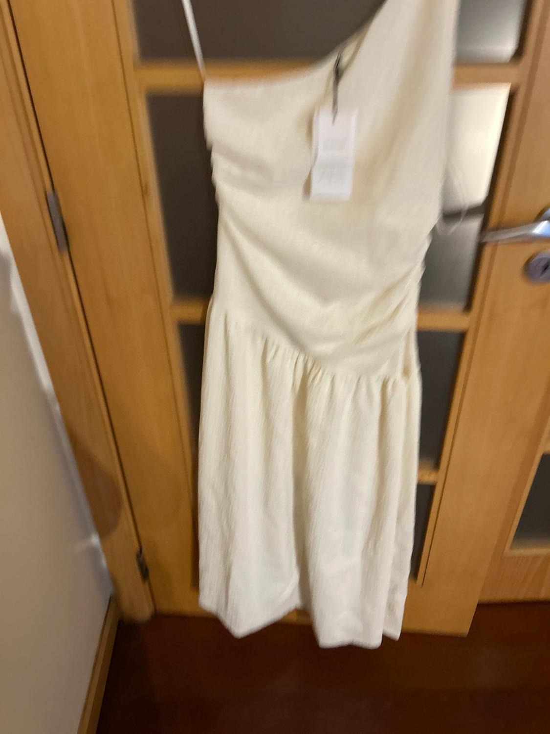 Asymmetrical white one-shoulder dress with ruching and a label, hanging on a wooden door with glass panels.