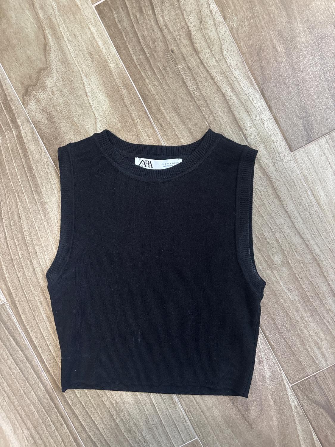 Black sleeveless top on light wooden floor. Visible tag with "ZARA" text.