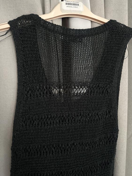Black openwork knit dress hanging on a beige hanger against a background of gray curtains. Visible tag: "putaway_induct".