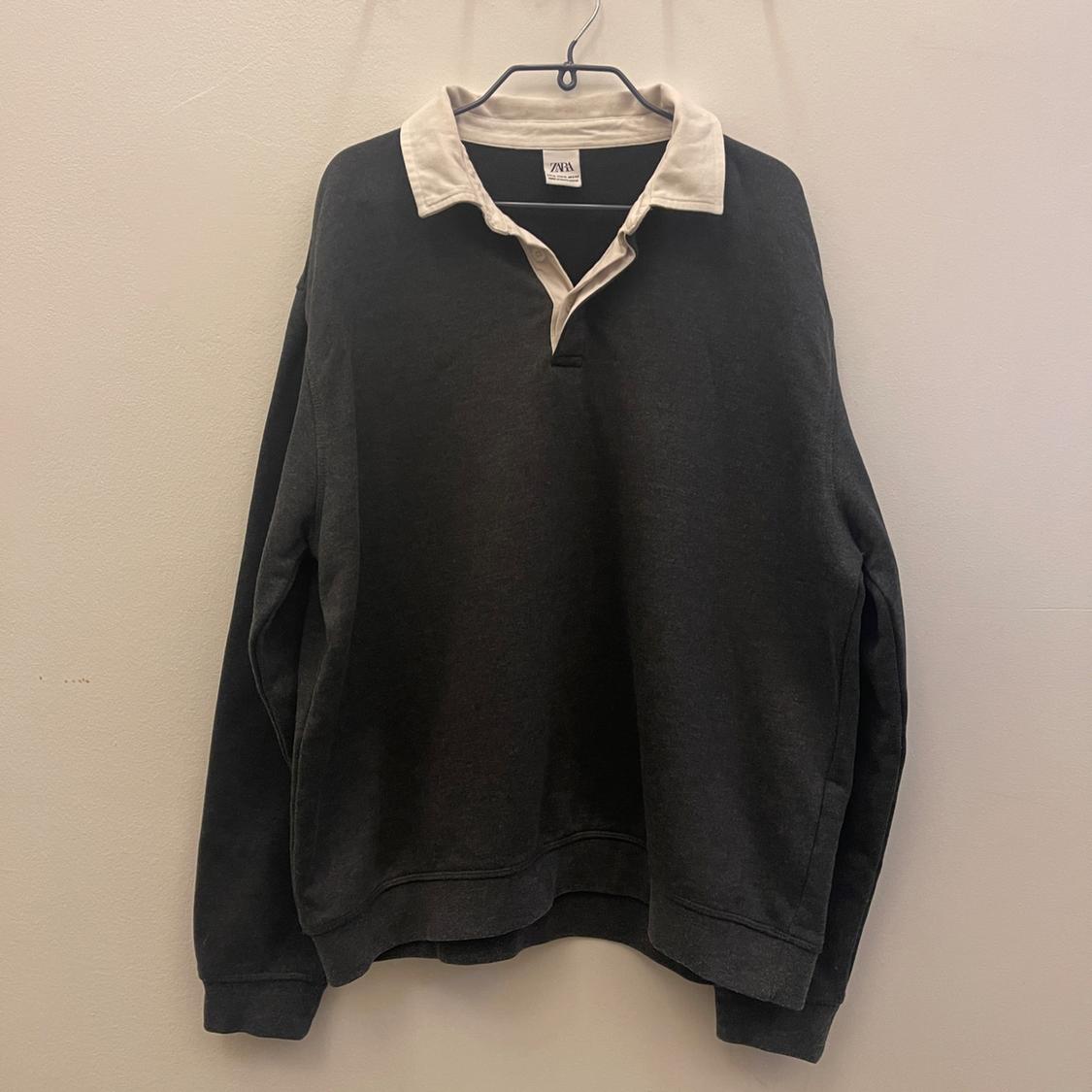 Black jumper with a white collar hanging on a hanger against a beige background. Label visible on the collar.