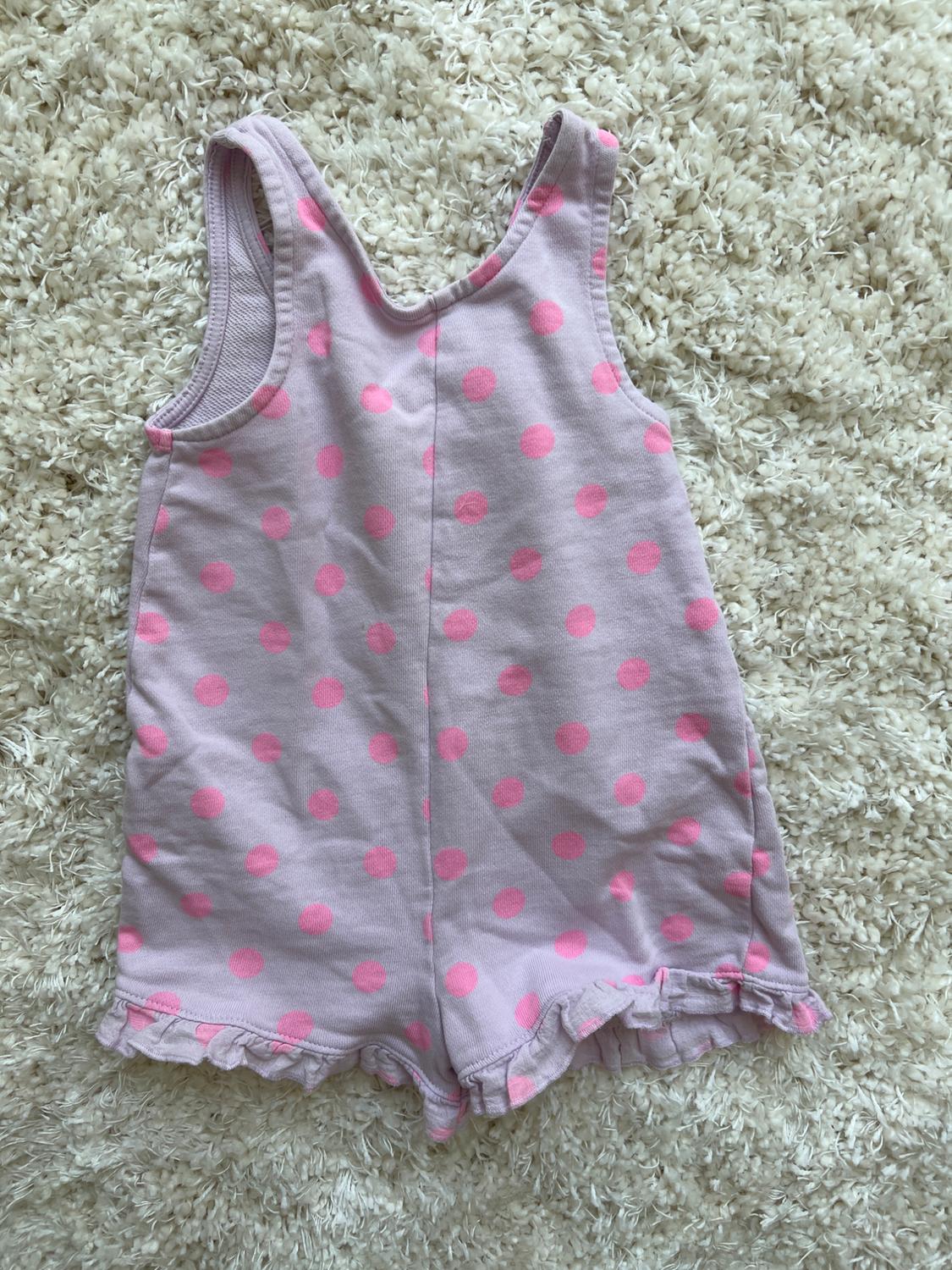 Pink children's all-in-one with fuchsia polka dots and ruffles on the hem, on a fluffy white textured rug.