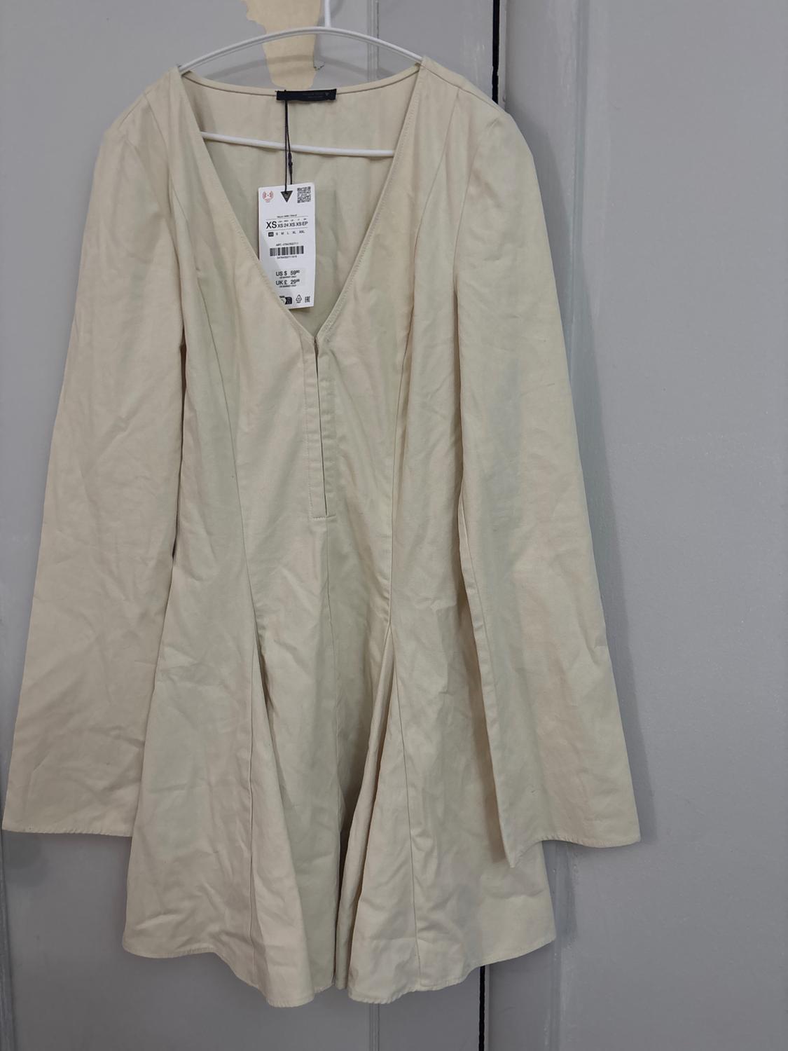 Beige dress with long, flared sleeves and a V-neckline, hanging on a hanger against a grey background. Visible label with the text XS.