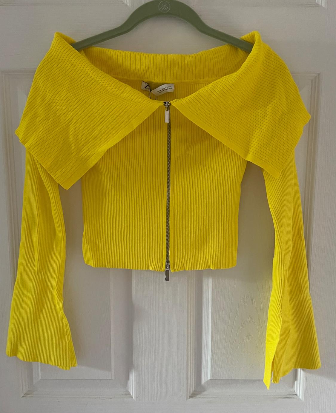 Yellow long-sleeved jacket with a front zip and a wide collar, hanging on a green hanger above a white door.