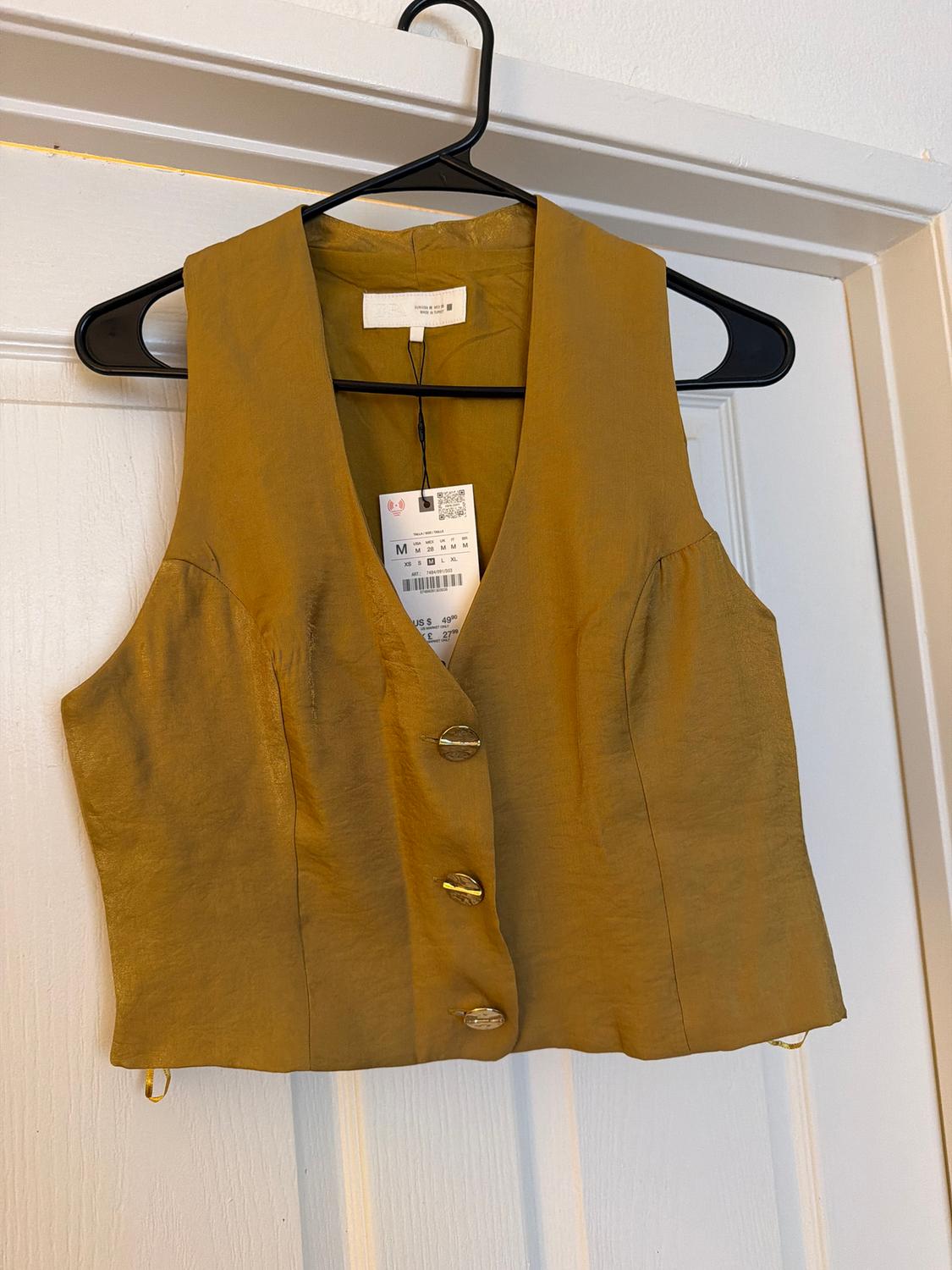Mustard sleeveless vest with large buttons, hanging on a black hanger over a white door. Tag visible.