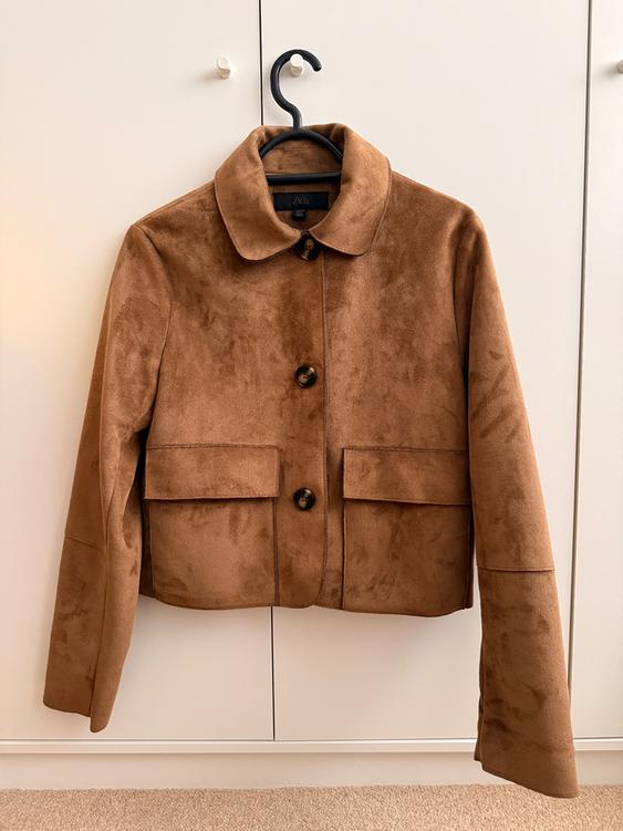 Brown suede jacket hanging on a black hanger in front of a white wardrobe, with two pockets and large buttons.