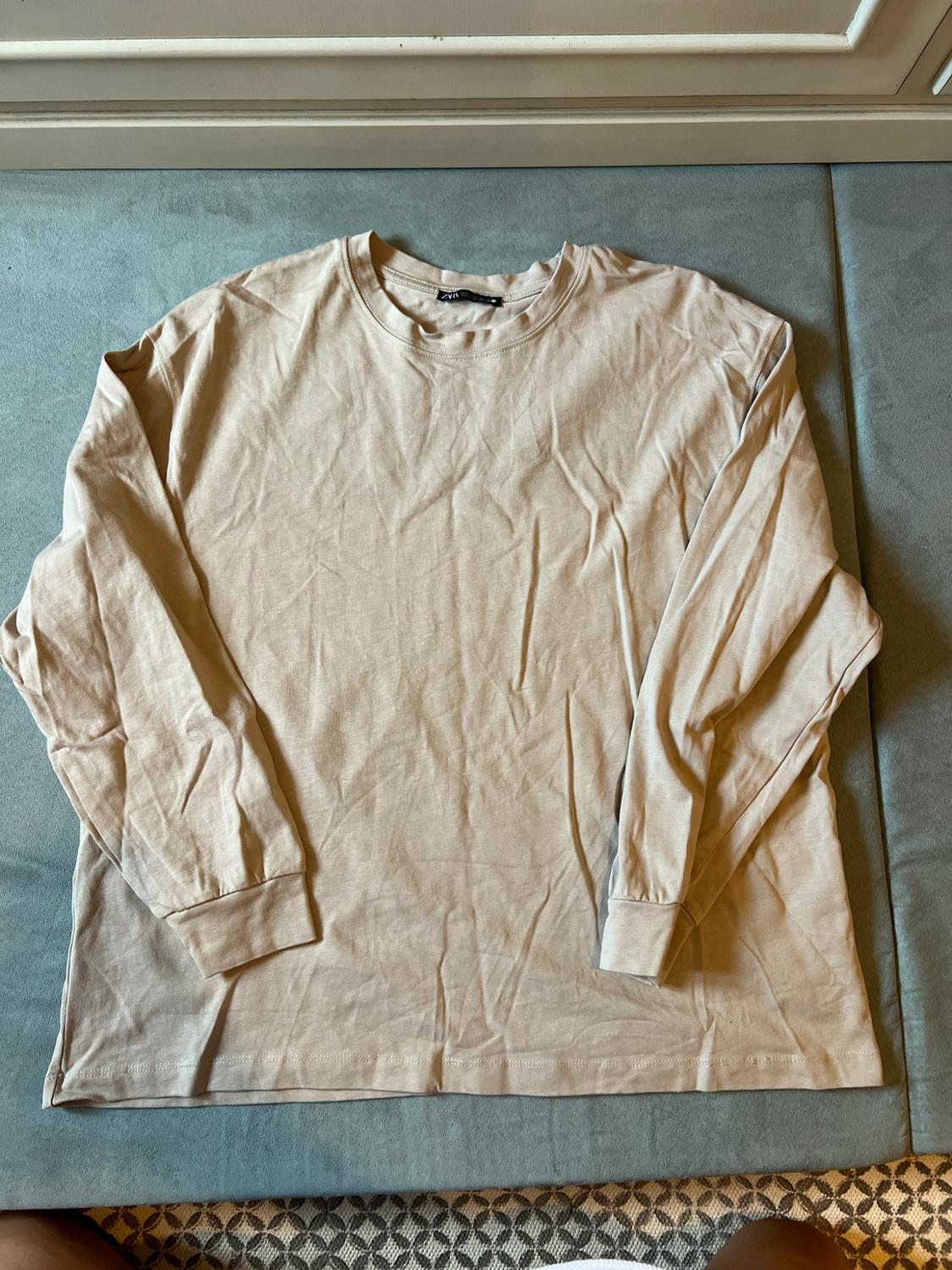 Light beige long-sleeved T-shirt laid out on a greyish-blue background. Interior label with the text ZARA.