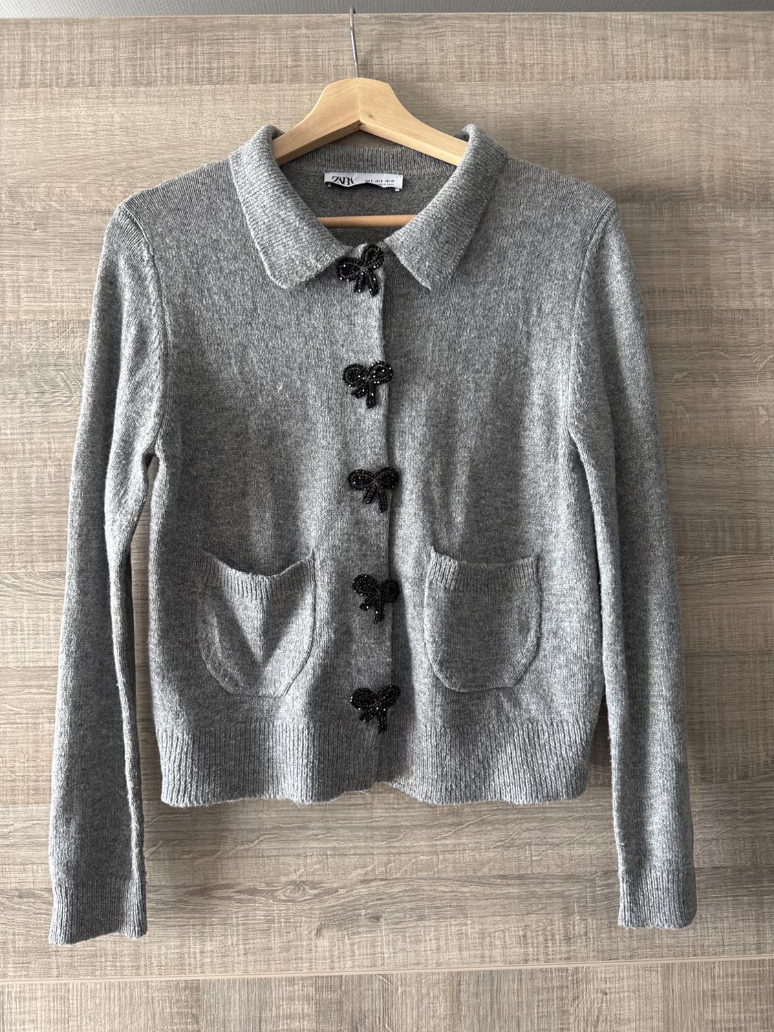 Grey knitted cardigan with a classic collar, decorative black buttons, and two front pockets, hanging on a wooden hanger.