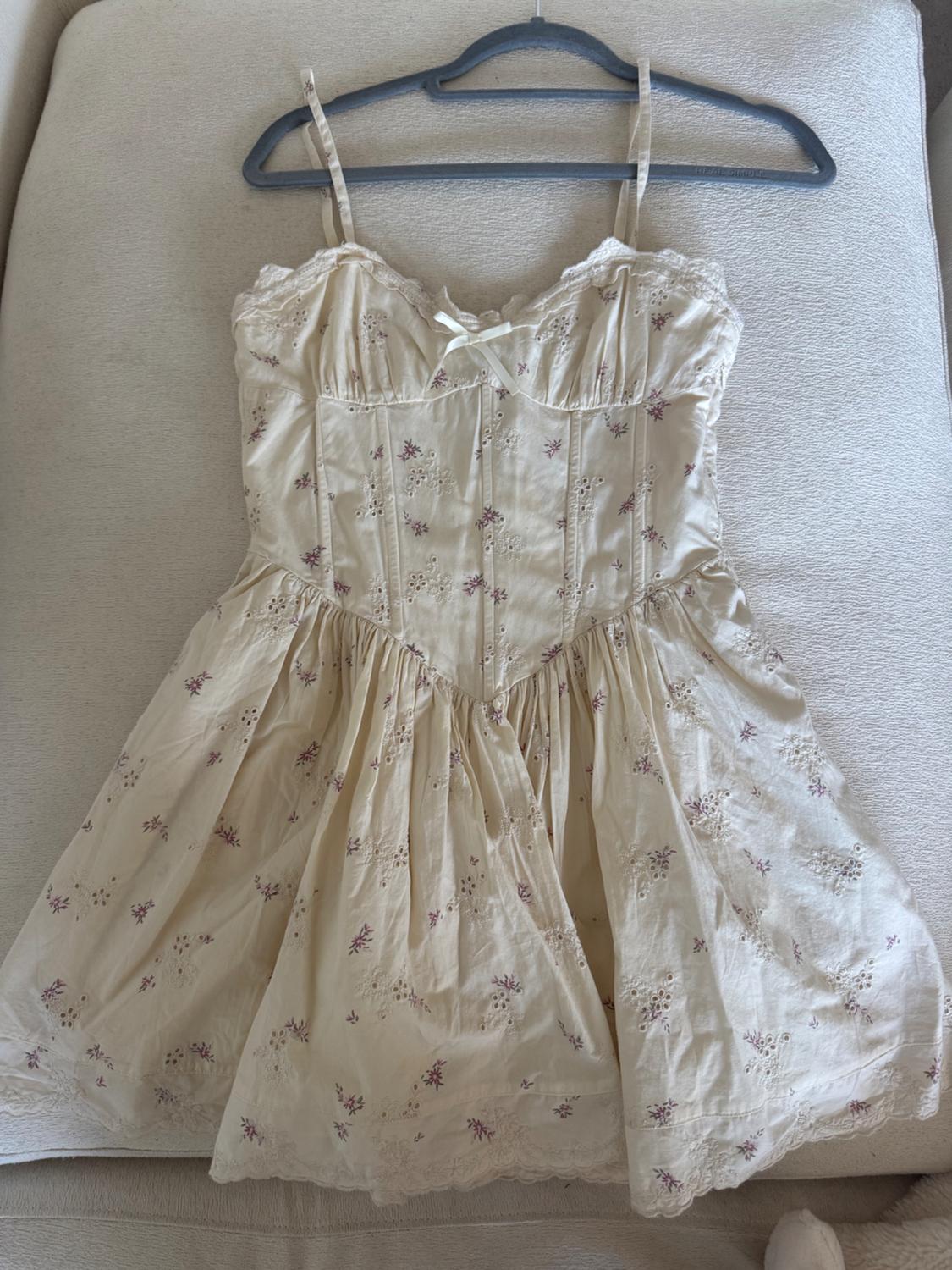 Cream dress with spaghetti straps and a lilac-toned floral print, hanging on a hanger over a beige sofa.