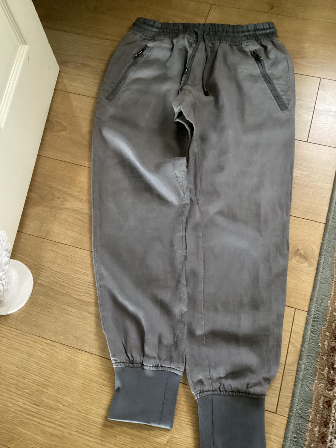 Grey joggers with an elasticated waistband and cuffs, drawstring and zip pockets, laid out on a wooden floor.