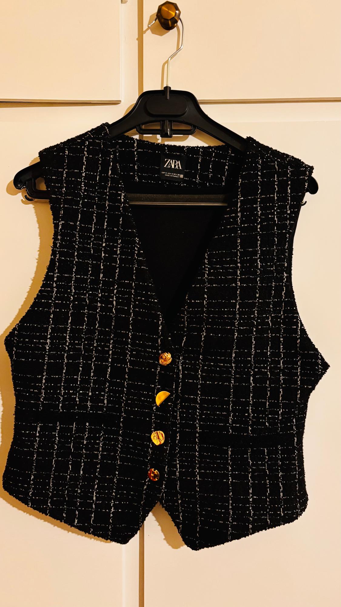 Black waistcoat with a white check pattern and gold buttons, hanging on a black hanger. Visible label: ZARA.