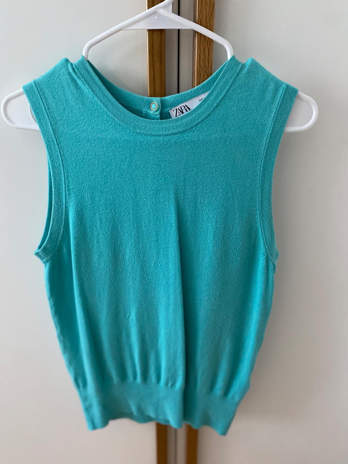 Turquoise blue sleeveless top hanging on a white hanger, with label visible on the neck. White and wood background.