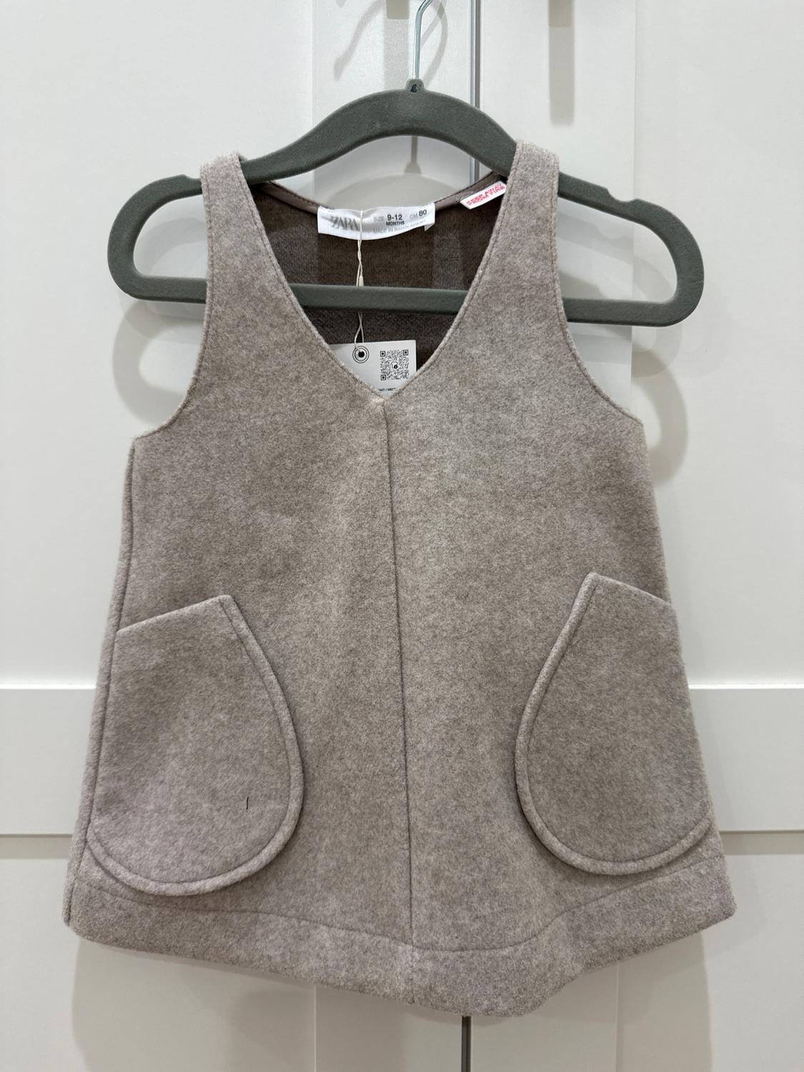 Sleeveless grey dress with large pockets, hanging on a black hanger. Visible tag with QR code and size 9-12 months.