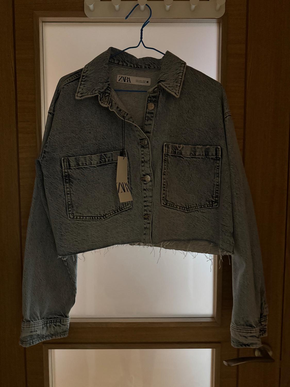 Cropped denim jacket hanging on a blue hanger in front of a glass door. Visible label with the text "ZARA".