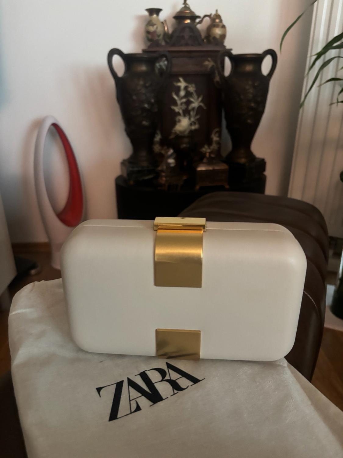 White hard-shell handbag with a gold clasp on a fabric bag bearing the text ZARA. Interior background with classic décor.