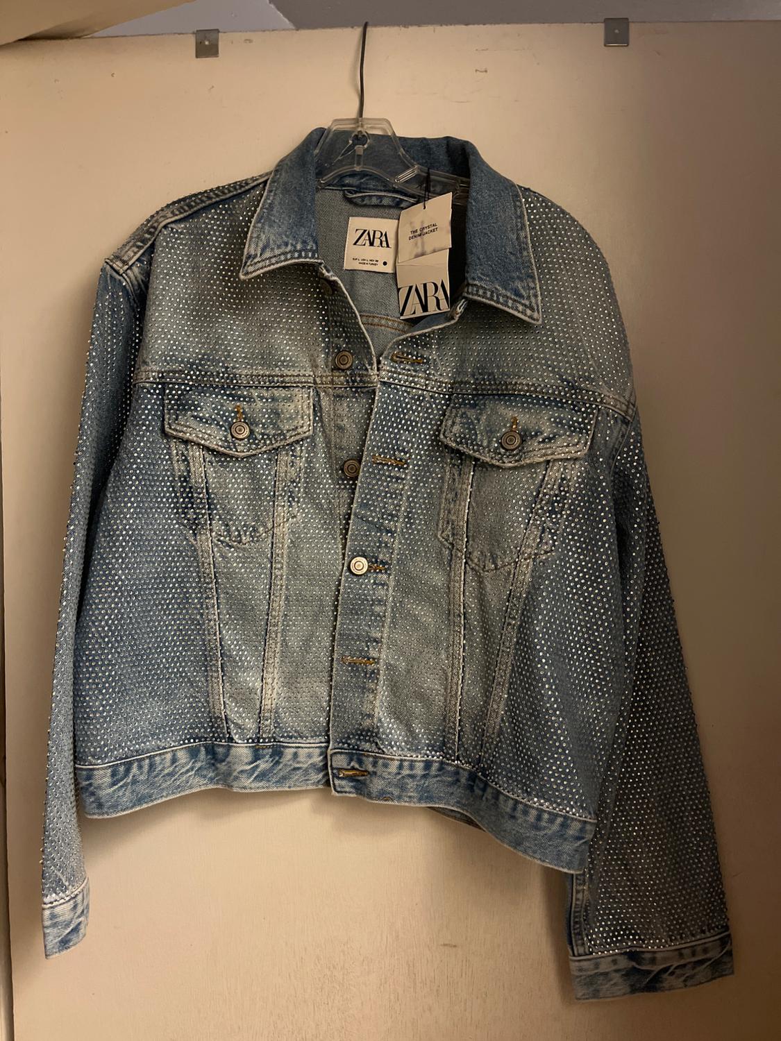 Blue denim jacket with silver studs hanging on a hanger. Zara labels visible on the collar. Beige background.