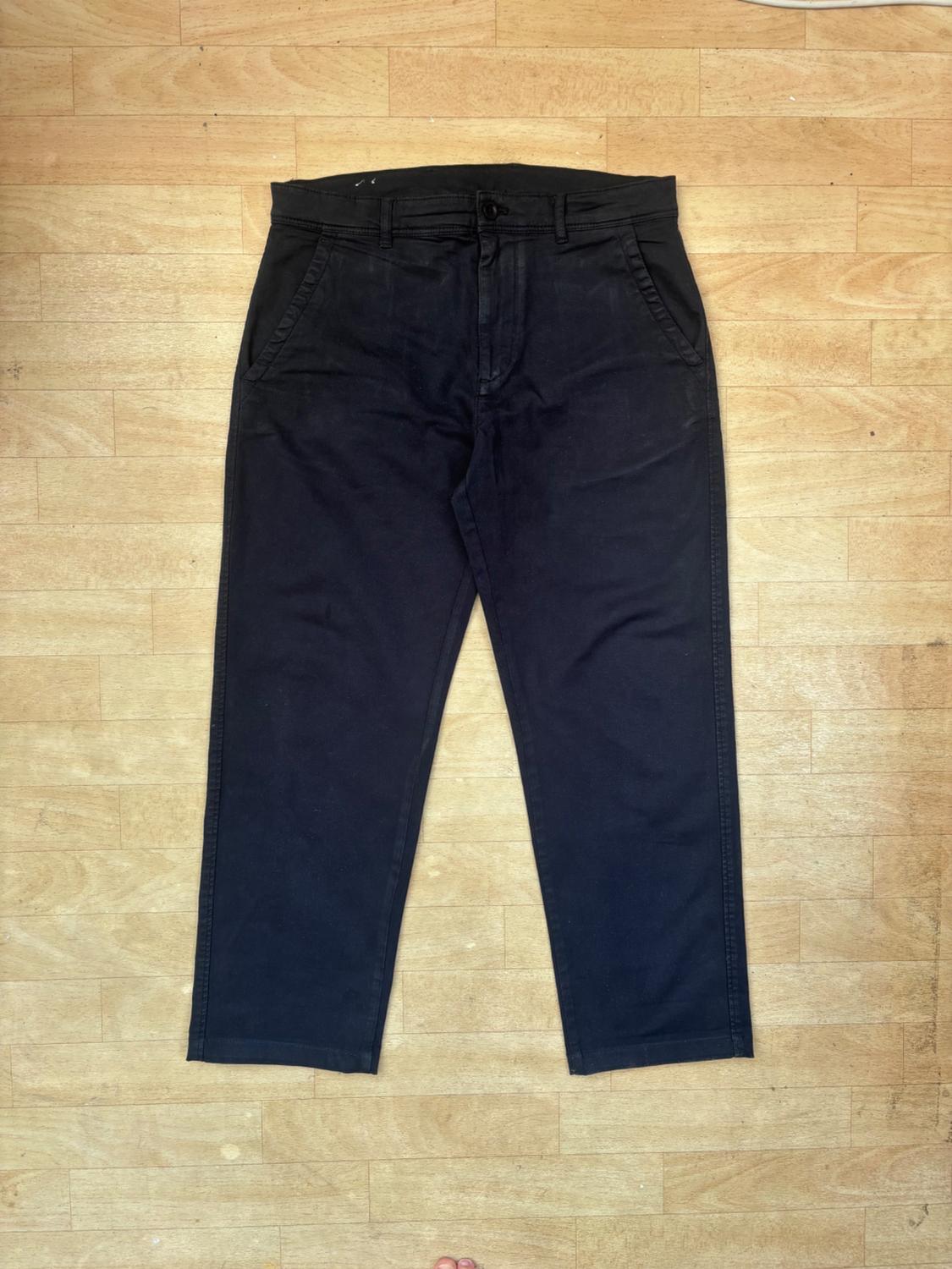 Dark casual trousers with pockets, belt loops and a button fastening, laid flat on a light wood floor.