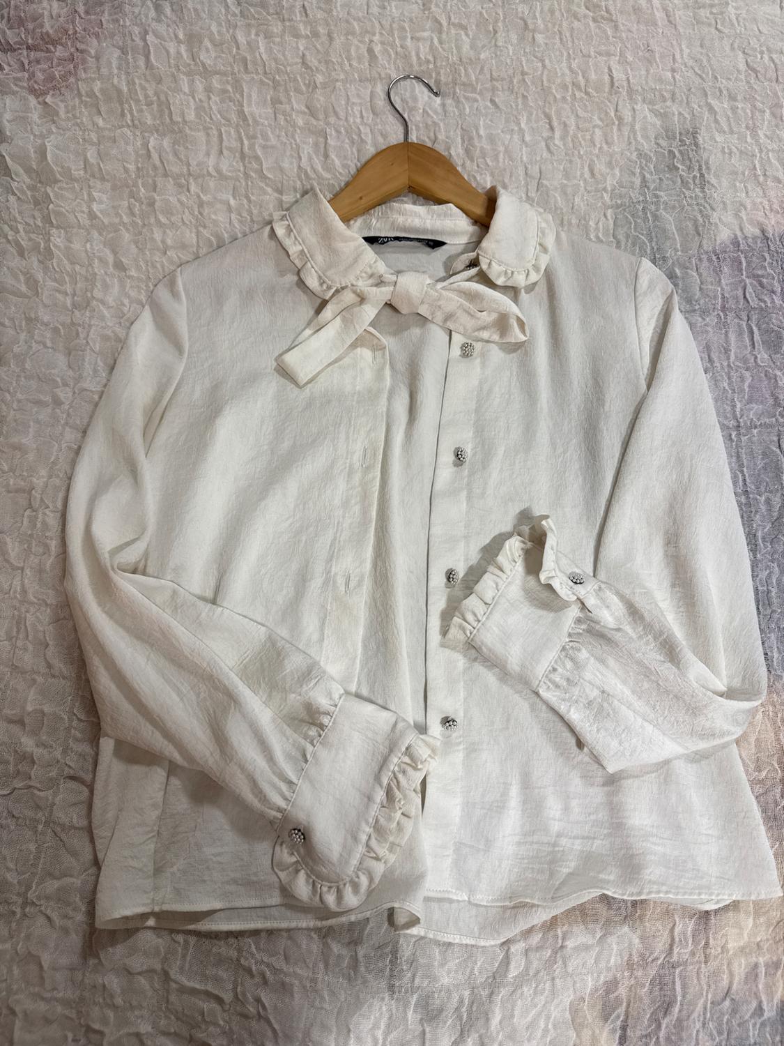 White blouse with ZARA label, textured, with a ruffled collar, tie detail and jewel buttons. On a wooden hanger against a crumpled light-coloured fabric.