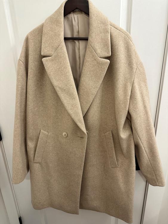 Beige wool coat hanging on a hanger, with wide lapels and two front pockets. White background of closet doors.