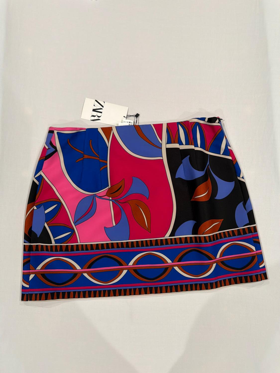 Short skirt on a white background with a vibrant print of leaves and geometric shapes in pink, blue, and black. Label with text ZARA.