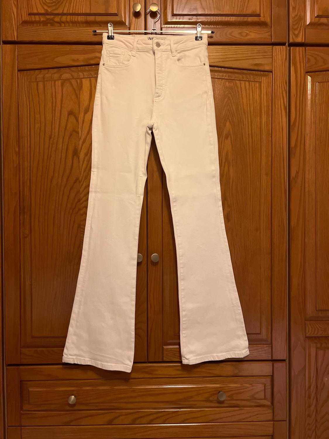 White high-waisted flared jeans, hanging on a clothes hanger in front of a wooden wardrobe.