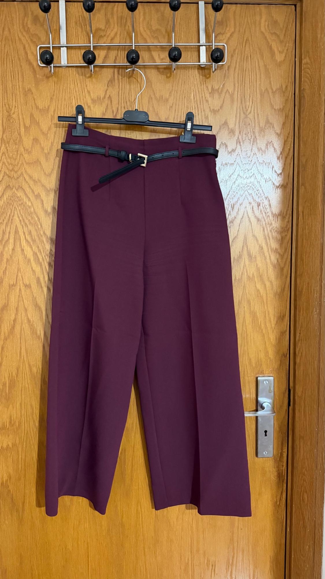 Burgundy wide-leg trousers with a black belt featuring a gold buckle, hanging on a hanger over a wooden door.