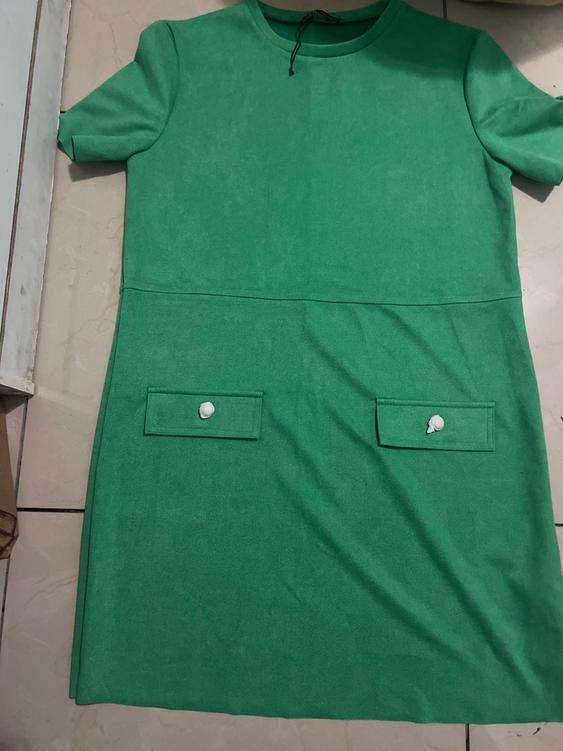 Short-sleeved green dress with two front pockets and white buttons, placed on a light-colored floor.